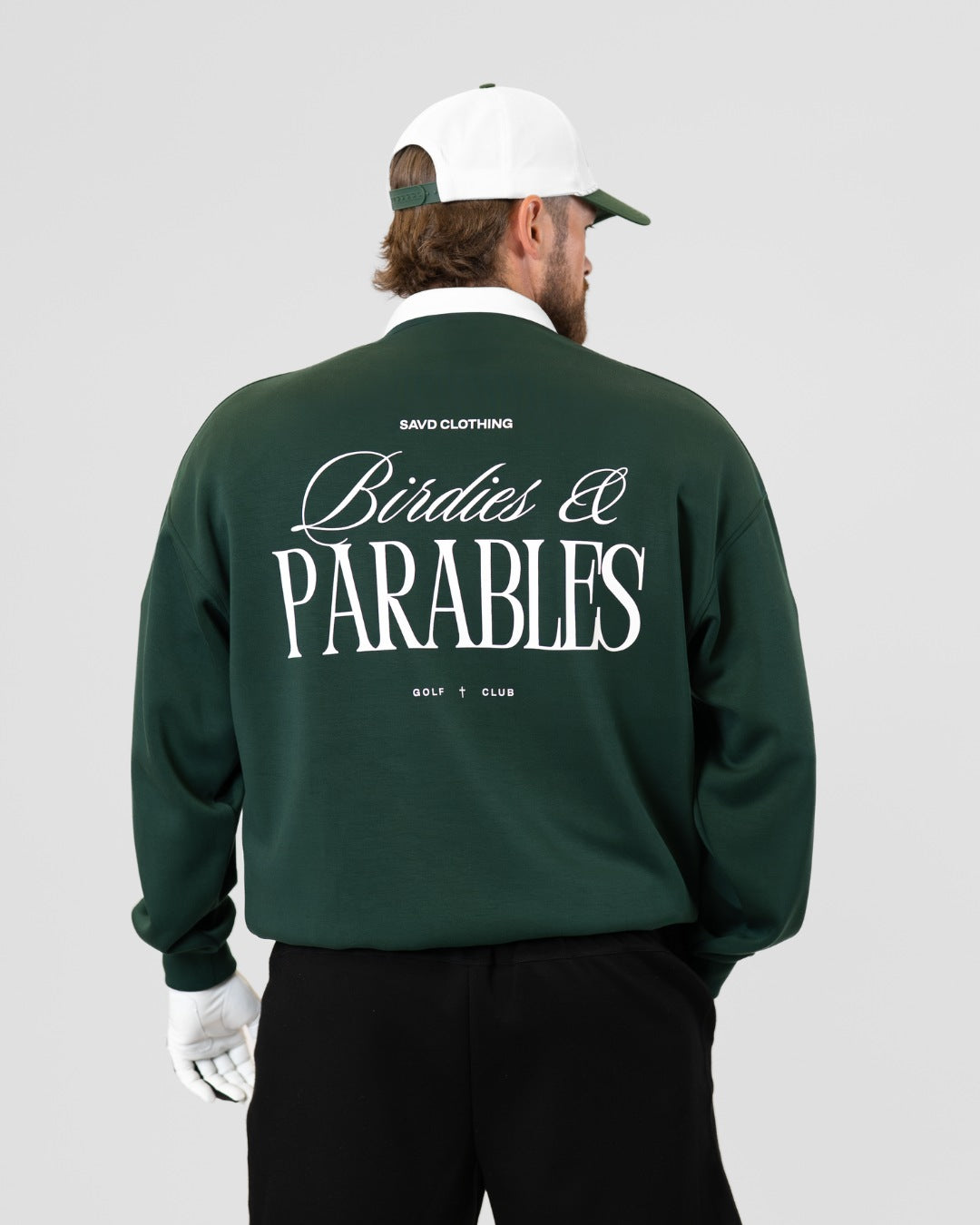 A man viewed from behind wearing a dark green sweatshirt with a large white graphic print reading 'Birdies & PARABLES' on the back, along with a white cap, white golf glove, and black pants, against a light gray studio background.