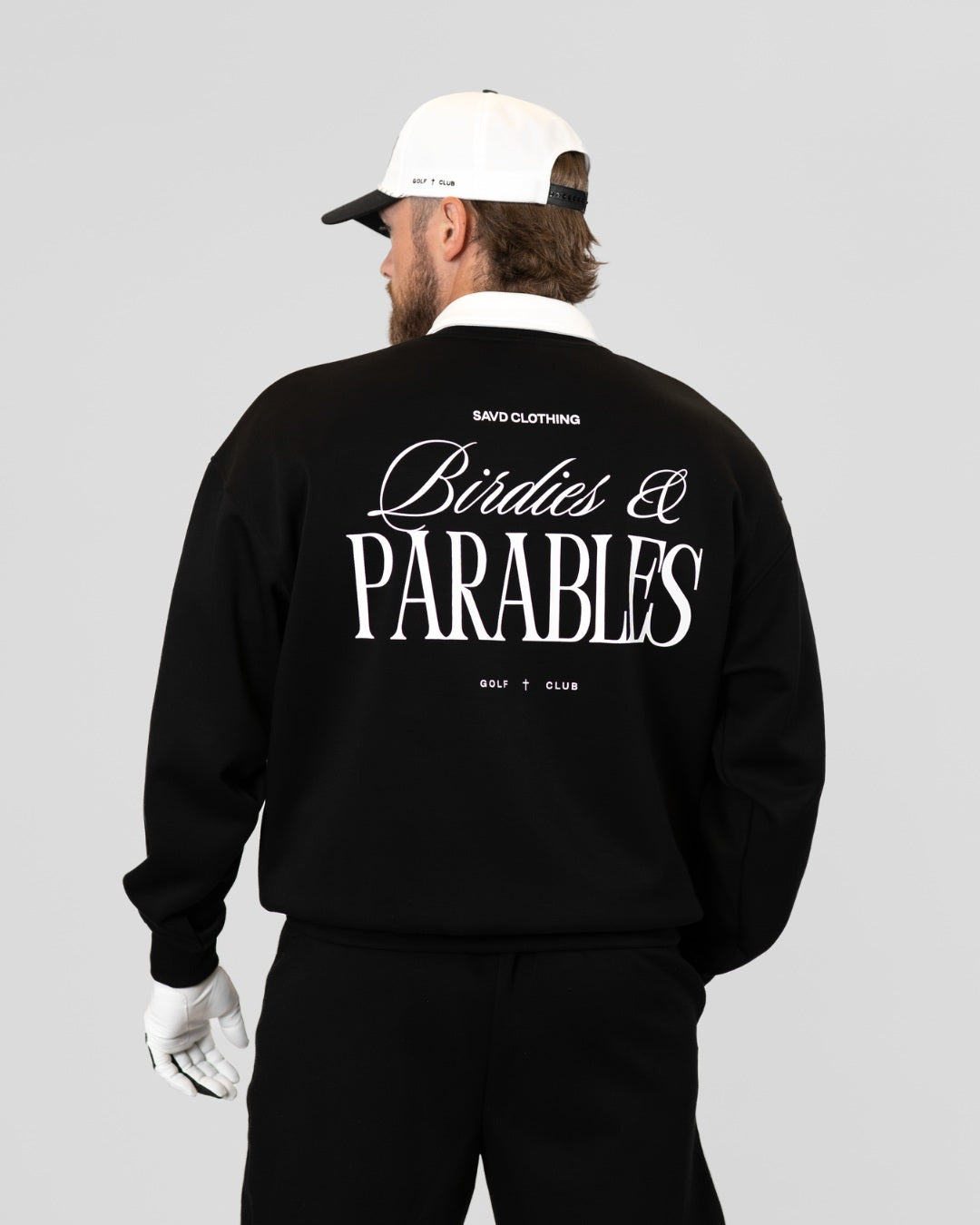 A man shown from the back wearing a black sweatshirt with large white 'Birdies & Parables' text on the back, a white cap, and black pants against a light gray studio background.