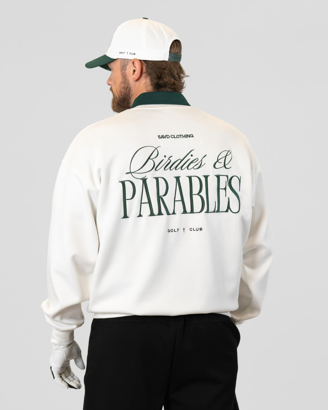 A man shown from the back wearing a white crewneck sweatshirt with a dark green collar and a large dark green graphic reading 'Birdies & PARABLES' on the back. He is also wearing a white and green baseball cap, a white golf glove, and black pants, set against a solid light gray background.